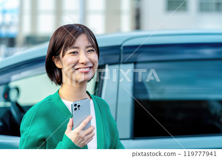 一名微笑的女士在汽車前拿著智慧型手機。 一名微笑的女士在汽車前拿著智慧型手機。 119777941