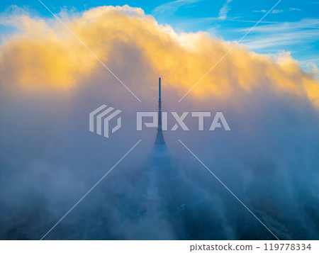 The towering structure stands amidst a dramatic, cloud-filled sunset, with vibrant hues illuminating the sky over Liberec, creating a captivating atmosphere. 119778334