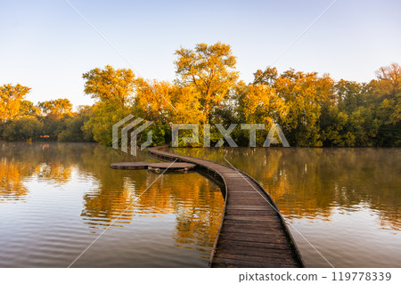 A narrow wooden bridge stretches over a calm pond, surrounded by vibrant autumn foliage on a peaceful morning in Vestec near Prague, creating a serene atmosphere. 119778339