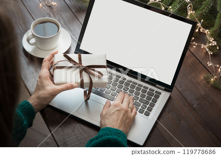 Christmas sales. Woman holding gift and typing by laptop in home interior. Screen as space for text. Xmas. Planning holidays. Christmas sales. Woman holding gift and typing by laptop in home interior. Screen as space for text. Xmas. Planning holidays. 119778686