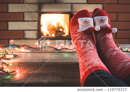 Feet in woollen red socks by the fireplace. Female relaxes by warm fire and warming up her feet in christmas socks. Close up on feet. Tabletop for display your christmas product. Christmas holiday. 119778727