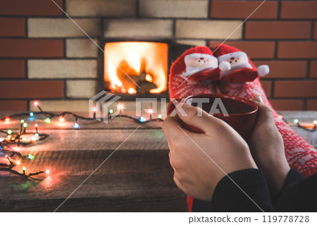 Female hands hold a red cup of coffee in front of the fireplace. Female relaxes by warm fire and warming up her feet in christmas red socks. Christmas holiday. 119778728