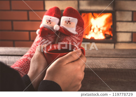 Teen's hands hold a red cup of coffee in front of the fireplace. Relax by warm fire and warming up her feet in christmas red socks. Christmas holiday. 119778729