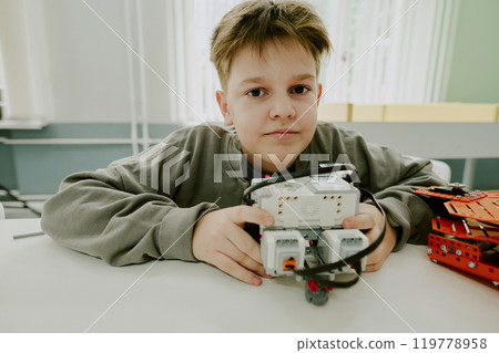 Medium close up portrait of adorable school boy cuddling prototype of robot while sitting at desk Medium close up portrait of adorable school boy cuddling prototype of robot while sitting at desk 119778958