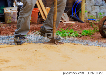 Worker uses manual rammer to tamp sand before constructing swimming pool 119780355