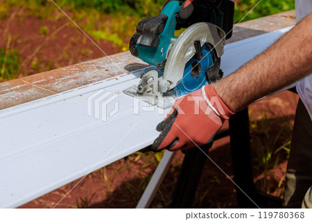 Prior to installing plastic vinyl siding, worker cuts it to size with circular handsaw 119780368