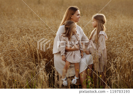Young woman with two daughters in a wheat field during sunset. 119783193