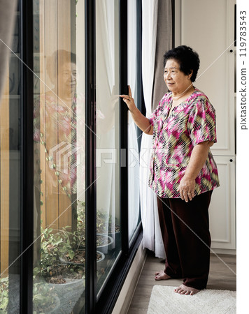 Asian Elderly woman standing by large window, touching glass, looking outside with thoughtful expression, wearing bright patterned shirt and brown pants, barefoot on carpet 119783543