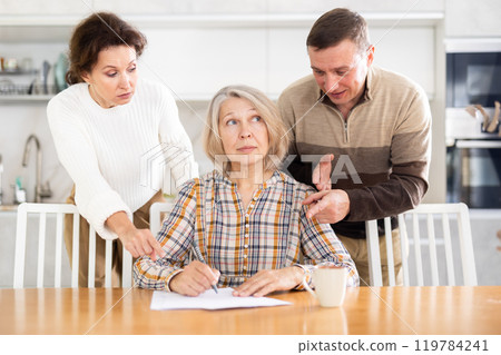 Old woman compiling heritage document while middle-aged man and woman competing in the kitchen 119784241