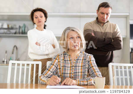 Old woman compiling heritage document while middle-aged man and woman competing in the kitchen 119784329