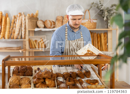 Man employee puts croissants in window, arranges display of goods at bakery. 119784363