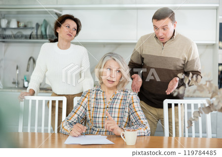 Old woman compiling heritage document while middle-aged man and woman competing in the kitchen 119784485