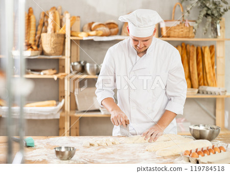 Baker in white uniform and cap cutting raw dough with knife to make croissants or buns 119784518