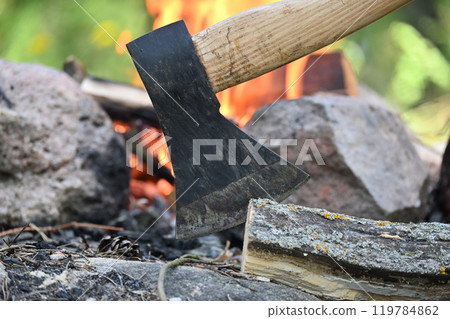 Hatchet lodged in a log of wood, situated opposite a campfire Hatchet lodged in a log of wood, situated opposite a campfire 119784862