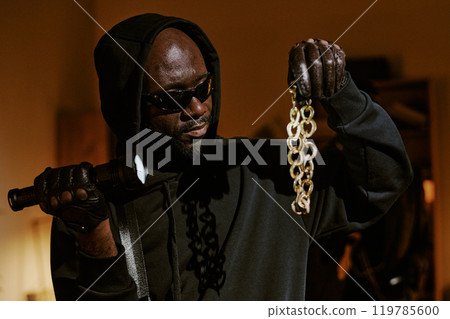 Medium close up of African American thief wearing hooded shirt and black glasses holding huge heavy golden chain 119785600