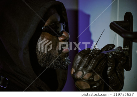 Medium close up of African American thief hiding his head under hood holding lockpick while trying to open door Medium close up of African American thief hiding his head under hood holding lockpick while trying to open door 119785627