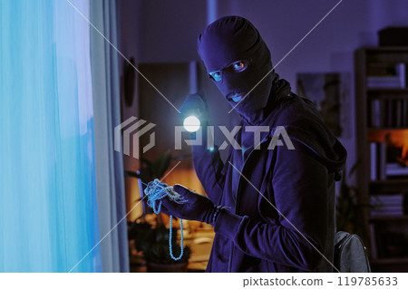Thief in balaclava and black clothes holding jewelry in hands and illuminating it with flash light while posing for portrait 119785633