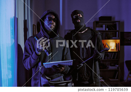 Two thieve in black clothes with crowbar, flashlight and stolen keyboard in hands hiding his face and head under hood and balaclava posing for portrait in dark room 119785639