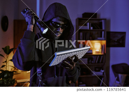 African American in total black clothes with hood looking at wireless keyboard in dark room African American in total black clothes with hood looking at wireless keyboard in dark room 119785641