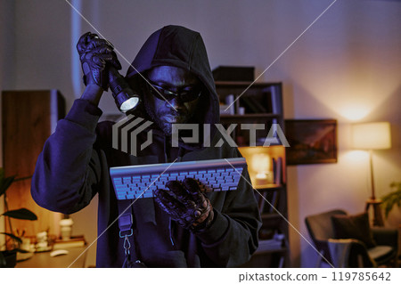African American thief illuminating wireless keyboard with flashlight while standing in dimly lit living room African American thief illuminating wireless keyboard with flashlight while standing in dimly lit living room 119785642