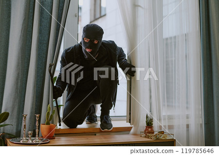 Man in total black with balaclava and crowbar sitting on windowsill after break-in 119785646