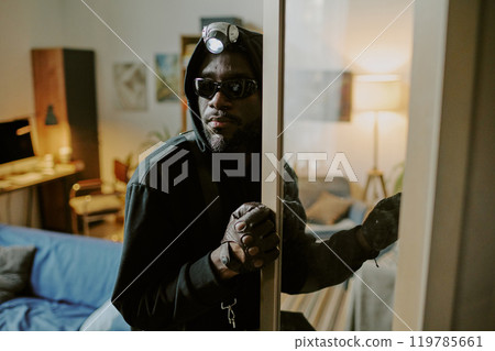 African American thief with headlamp opening cupboard carefully while glancing around 119785661