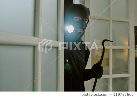 Thief with flashlight and metal crowbar glancing around apartment 119785663