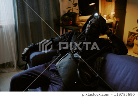 African American thief in black clothes and leather gloves sitting on sofa relaxed during trespassing African American thief in black clothes and leather gloves sitting on sofa relaxed during trespassing 119785690