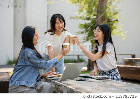 A group of cheerful Asian college students is enjoying coffee together at a table in a college park. A group of cheerful Asian college students is enjoying coffee together at a table in a college park. 119785729