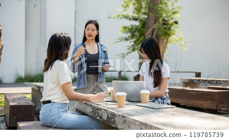 A smart Asian female college student is tutoring math to her friends at a table in a college park. A smart Asian female college student is tutoring math to her friends at a table in a college park. 119785739