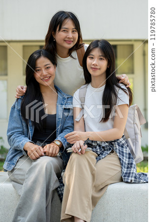 A group of Asian students is sitting on a bench in their college park, smiling at the camera. 119785780