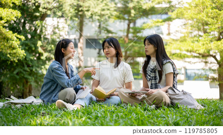 A group of college students sits on the grass in their college campus park, having a conversation. 119785805