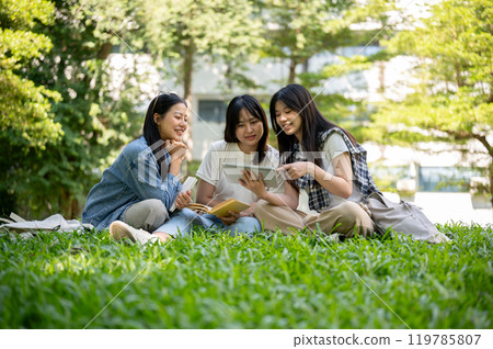 A group of students sits on the grass, chatting about a video they're watching together on a tablet. 119785807