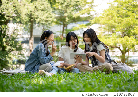 A group of students sits on the grass, chatting about a video they're watching together on a tablet. 119785808