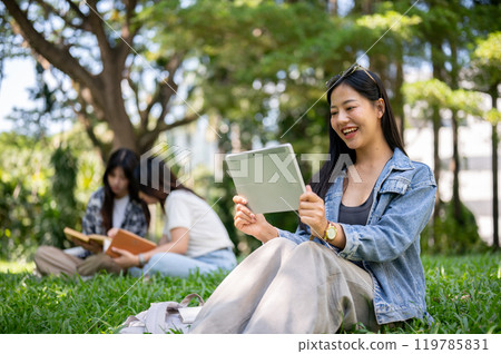 A beautiful Asian female college student sits on grass in a park, using her digital tablet. 119785831