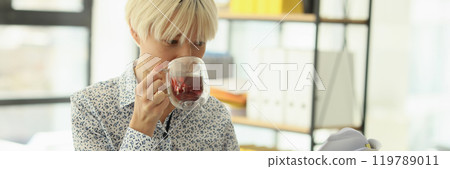 Blonde woman drinks tea in glass cup reading document 119789011