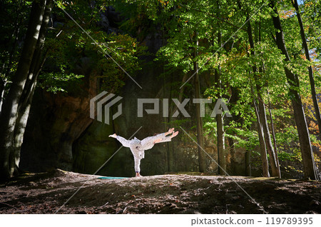 Woman practicing yoga outdoors in forest. Barefoot female on yoga mat surrounded by trees and large rocks, which suggests peaceful, natural environment ideal for meditation or yoga practice. 119789395