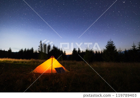 Glowing orange tent illuminated at dusk beside solar panel in grassy field. Backdrop of silhouetted trees and starry night sky emphasizes eco-friendly and serene camping experience. 119789403