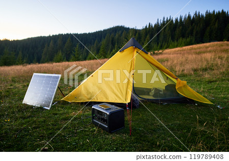 Tourist tent with photovoltaic solar panel and portable power station nearby in grassy field at sunset. Scene surrounded by rolling hills under clear blue sky, showcasing eco-friendly camping setup. 119789408