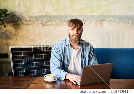 Bearded man works on laptop at wooden table, with cup of coffee. Solar panel nearby, emphasizing sustainable eco-friendly workspace that combines technology and sustainability in cozy environment. 119789414