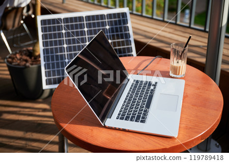 Laptop on round wooden table next to glass of iced coffee on sunny balcony. In background, solar panel propped up, symbolizing blend of technology and sustainable energy in modern workspace. 119789418