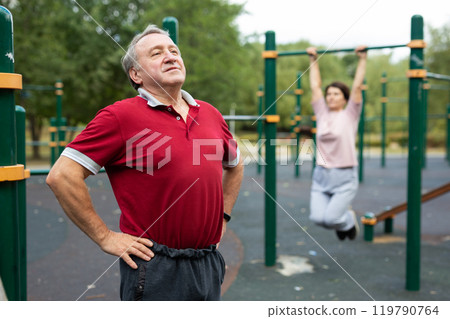 Staying after retirement. Happy joyful mature retired sportsman wearing sportswear doing side stretching exercises with arm over his head, exercising outside in city park Staying after retirement. Happy joyful mature retired sportsman wearing sportswear doing side stretching exercises with arm over his head, exercising outside in city park 119790764