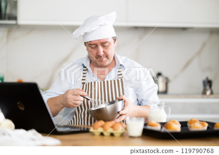 Man in a chef's hat and apron prepares cupcakes in the kitchen Man in a chef's hat and apron prepares cupcakes in the kitchen 119790768