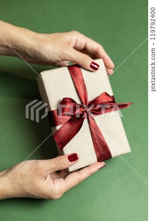a female hands with a burgundy manicure holds a gift box with a burgundy bow a female hands with a burgundy manicure holds a gift box with a burgundy bow 119792400