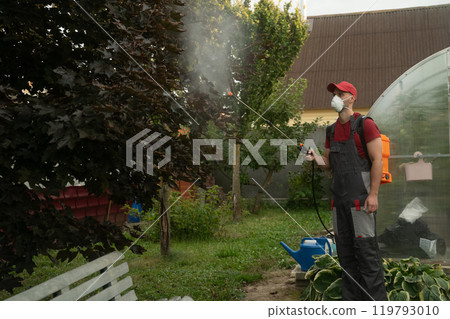 Gardener wearing mask spraying trees in garden 119793010