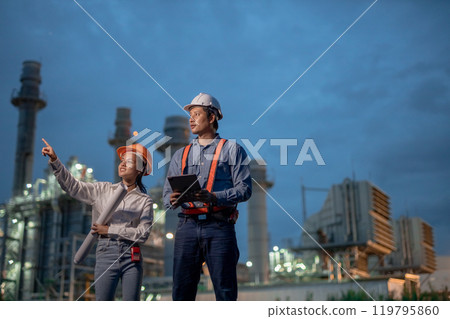 Asian team man and women engineer working at nighttime with blueprint standing at front of Oil-fired power plant. Worker wear uniform helmet working tower power plant construction. 119795860