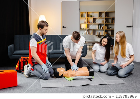 Young man instructor helping to make first aid heart compressions with dummy during the group training indoors Young man instructor helping to make first aid heart compressions with dummy during the group training indoors 119795969