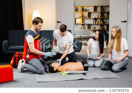 Young man instructor helping to make first aid heart compressions with dummy during the group training indoors Young man instructor helping to make first aid heart compressions with dummy during the group training indoors 119795970