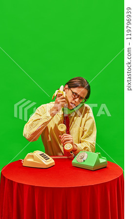 Man humorously holds banana as phone receiver and try to eat receiver while sitting at table, combining humor and vintage office elements. Man humorously holds banana as phone receiver and try to eat receiver while sitting at table, combining humor and vintage office elements. 119796939