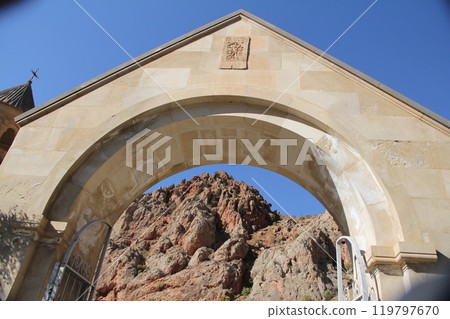 Stone archway entrance with a carved relief, set against a mountainous landscape and clear blue sky. 119797670